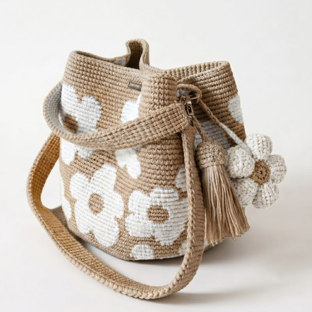 Borsa a secchiello all'uncinetto fatta a mano, di colore beige con un motivo di grandi fiori bianchi, posizionata su uno sfondo bianco. La borsa è completa di una tracolla intrecciata beige, due nappine e un charm a forma di fiore all'uncinetto.