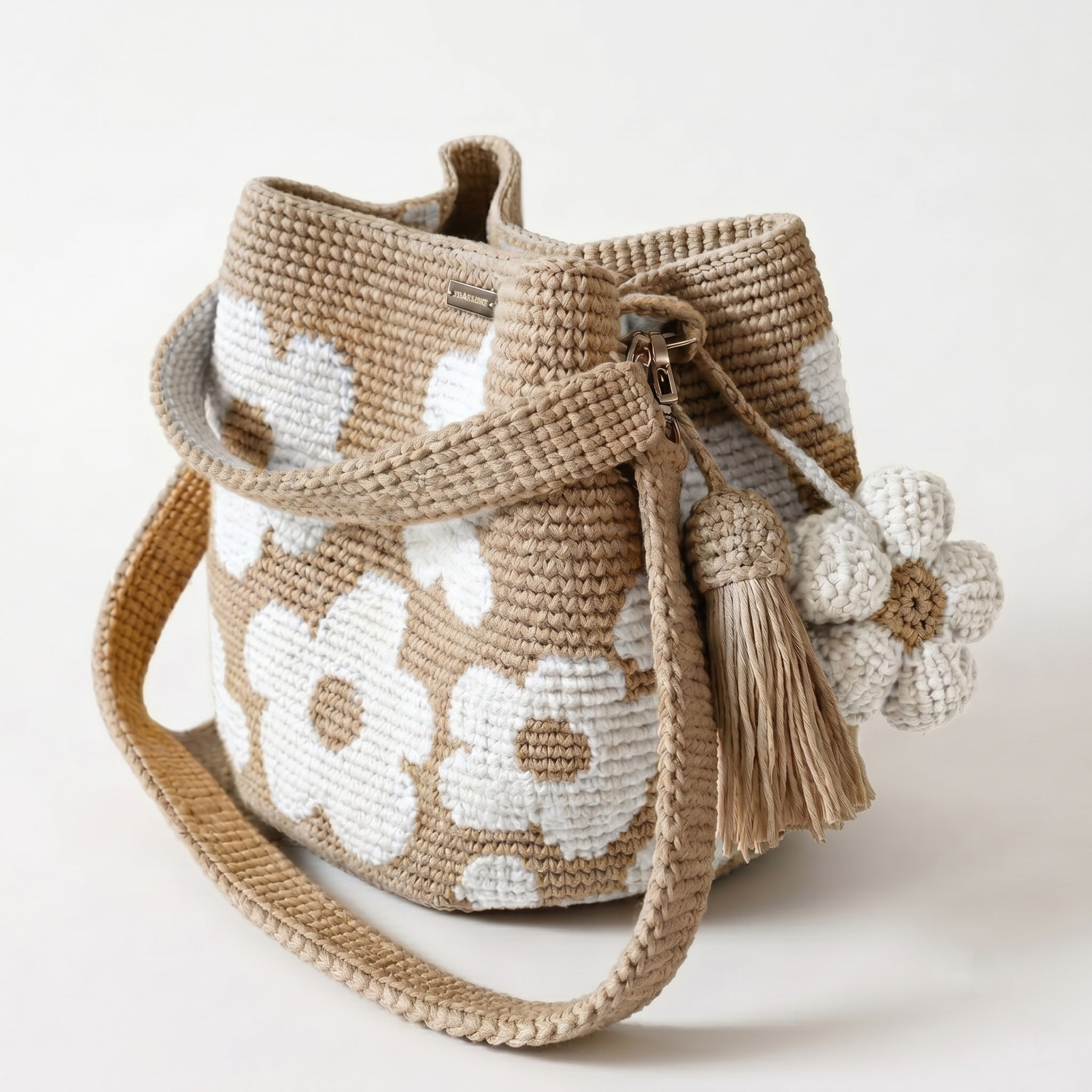 Borsa a secchiello all'uncinetto fatta a mano, di colore beige con un motivo di grandi fiori bianchi, posizionata su uno sfondo bianco. La borsa è completa di una tracolla intrecciata beige, due nappine e un charm a forma di fiore all'uncinetto.