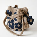 Borsa a secchiello all'uncinetto fatta a mano, di colore beige con un motivo di grandi fiori blu, posizionata su uno sfondo bianco. La borsa è completa di una tracolla intrecciata beige, due nappine e un charm a forma di fiore all'uncinetto.
