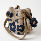 Borsa a secchiello all'uncinetto fatta a mano, di colore beige con un motivo di grandi fiori blu, posizionata su uno sfondo bianco. La borsa è completa di una tracolla intrecciata beige, due nappine e un charm a forma di fiore all'uncinetto.