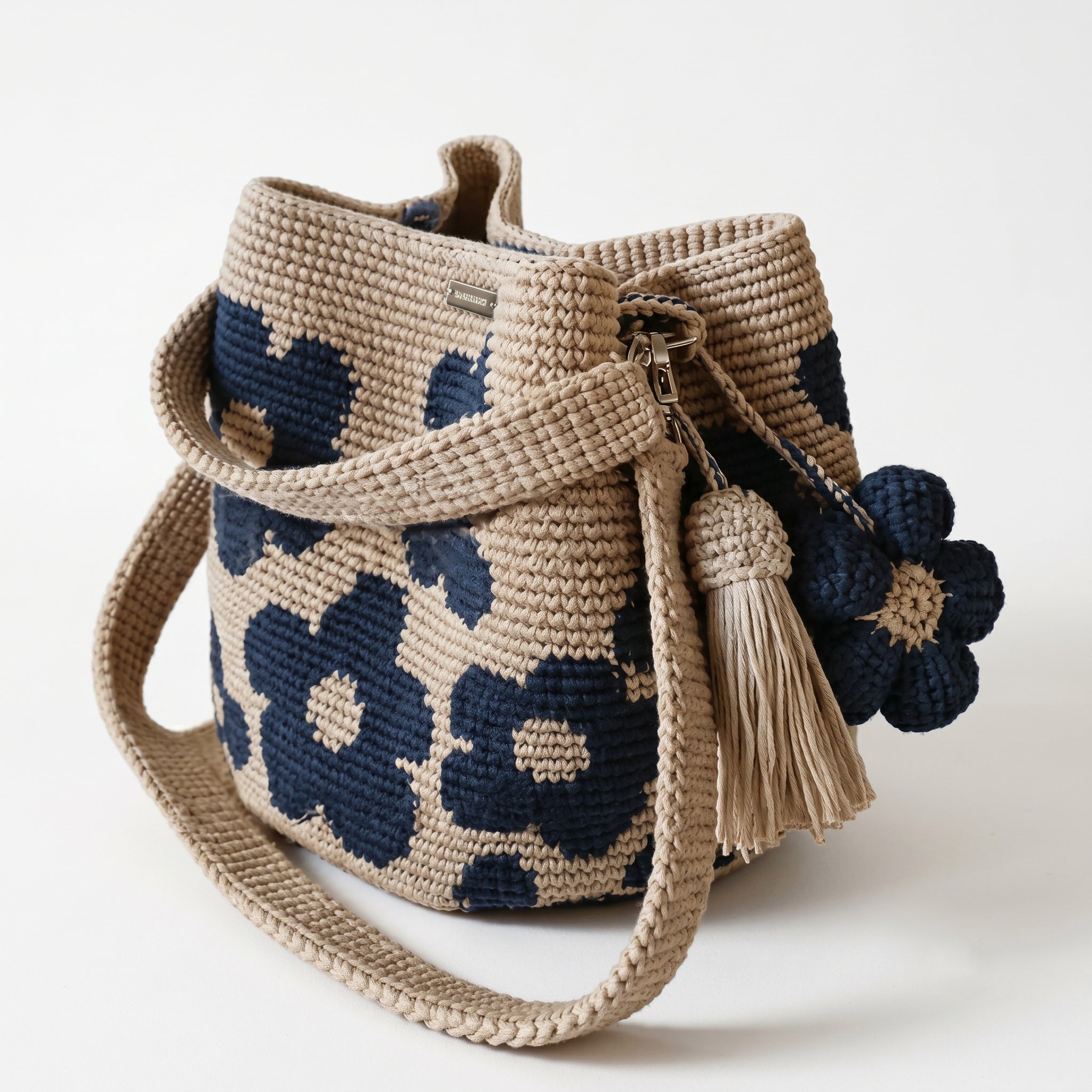 Borsa a secchiello all'uncinetto fatta a mano, di colore beige con un motivo di grandi fiori blu, posizionata su uno sfondo bianco. La borsa è completa di una tracolla intrecciata beige, due nappine e un charm a forma di fiore all'uncinetto.