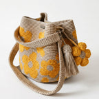 Borsa a secchiello all'uncinetto fatta a mano, di colore beige con un motivo di grandi fiori gialli, posizionata su uno sfondo bianco. La borsa è completa di una tracolla intrecciata beige, due nappine e un charm a forma di fiore all'uncinetto.