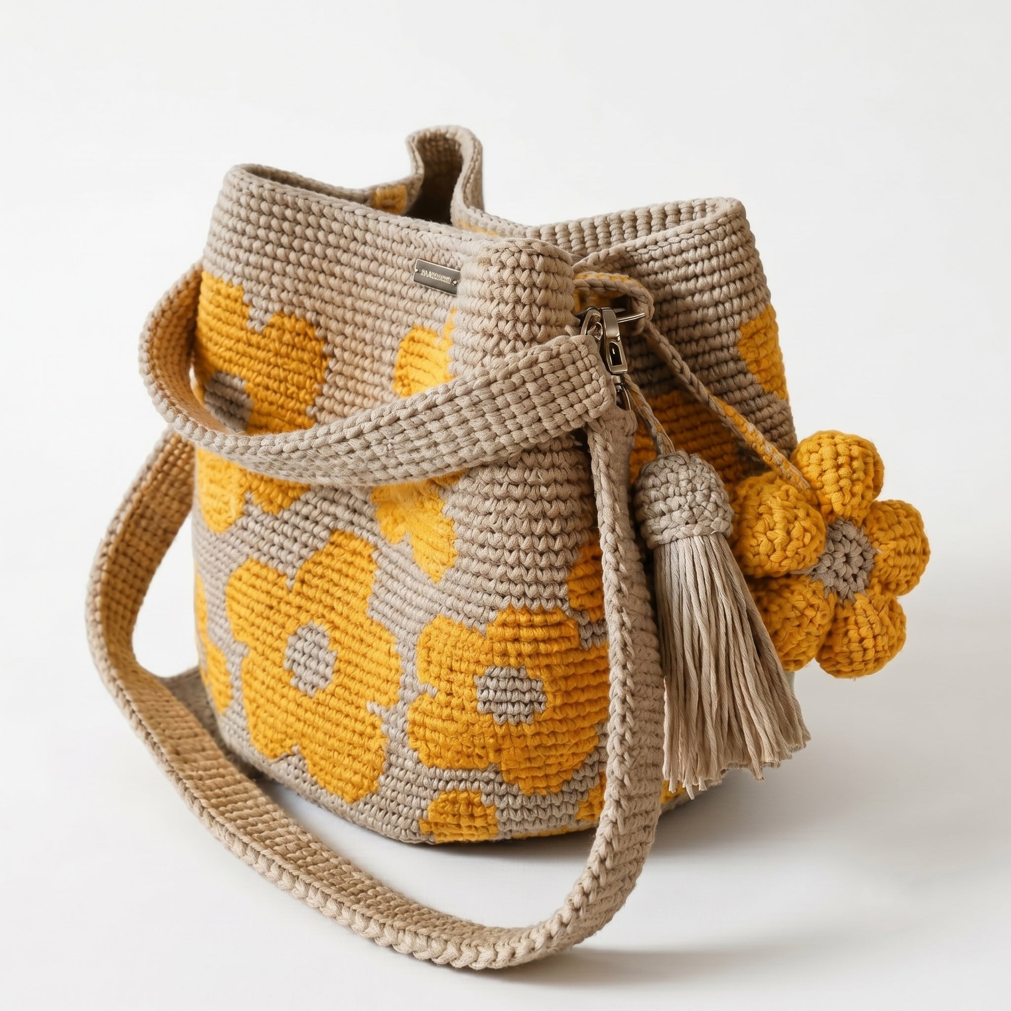 Borsa a secchiello all'uncinetto fatta a mano, di colore beige con un motivo di grandi fiori gialli, posizionata su uno sfondo bianco. La borsa è completa di una tracolla intrecciata beige, due nappine e un charm a forma di fiore all'uncinetto.
