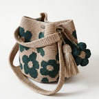 Borsa a secchiello all'uncinetto fatta a mano, di colore beige con un motivo di grandi fiori verdi, posizionata su uno sfondo bianco. La borsa è completa di una tracolla intrecciata beige, due nappine e un charm a forma di fiore all'uncinetto.