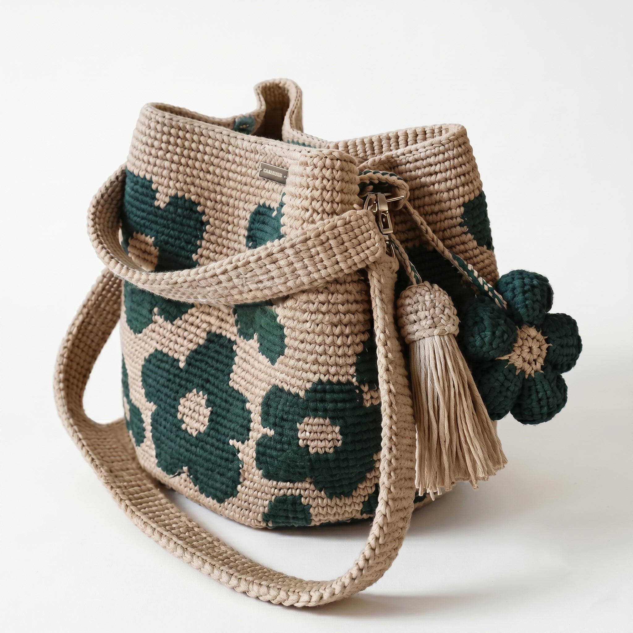 Borsa a secchiello all'uncinetto fatta a mano, di colore beige con un motivo di grandi fiori verdi, posizionata su uno sfondo bianco. La borsa è completa di una tracolla intrecciata beige, due nappine e un charm a forma di fiore all'uncinetto.