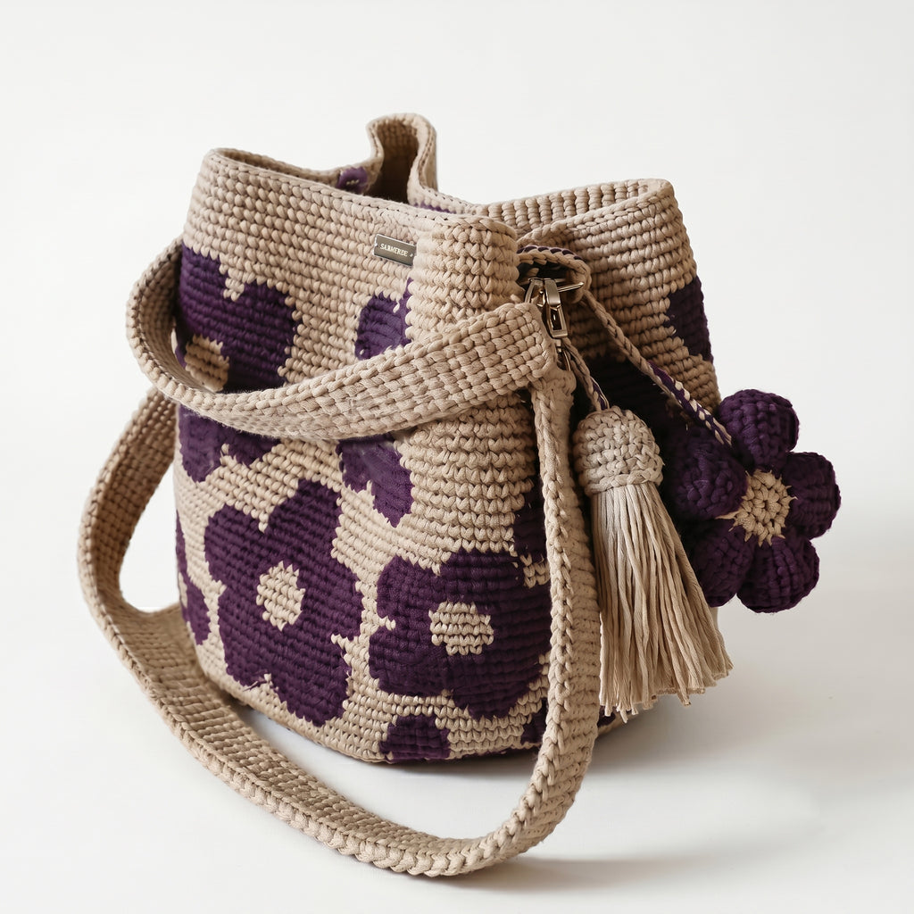 Borsa a secchiello all'uncinetto fatta a mano, di colore beige con un motivo di grandi fiori viola, posizionata su uno sfondo bianco. La borsa è completa di una tracolla intrecciata beige, due nappine e un charm a forma di fiore all'uncinetto.