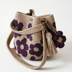 Borsa a secchiello all'uncinetto fatta a mano, di colore beige con un motivo di grandi fiori viola, posizionata su uno sfondo bianco. La borsa è completa di una tracolla intrecciata beige, due nappine e un charm a forma di fiore all'uncinetto.